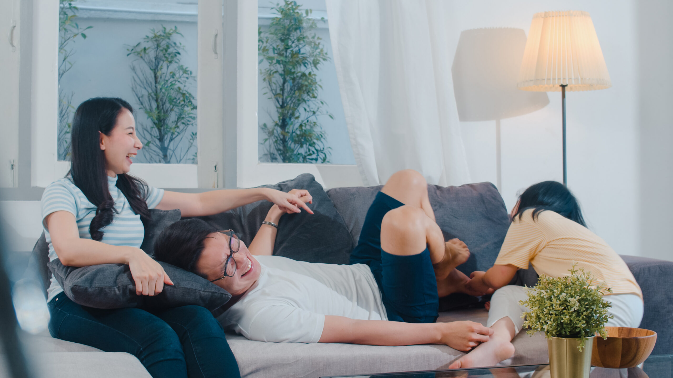 Happy young Asian family play together on couch at home. Chinese mother father and child daughter enjoying happy relax spending time together in modern living room in evening.